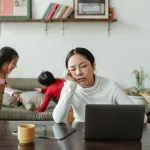 A tired mother working remotely on her laptop while children play in the background.